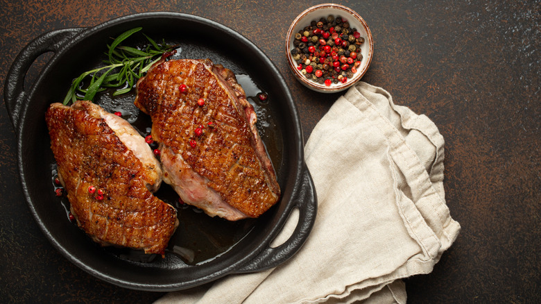 Two duck breasts in cast iron pan next to bowl of peppercorns and a dish towel