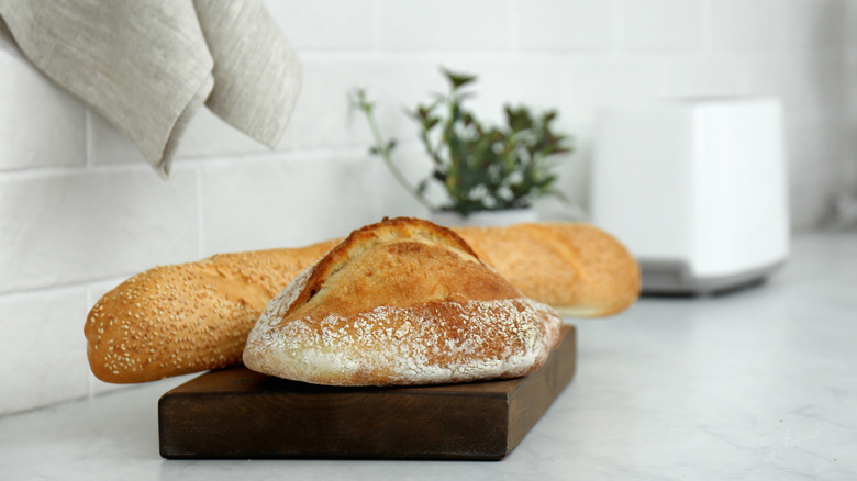 A loaf of bread and a baguette sit on a dark wood cutting board on counter