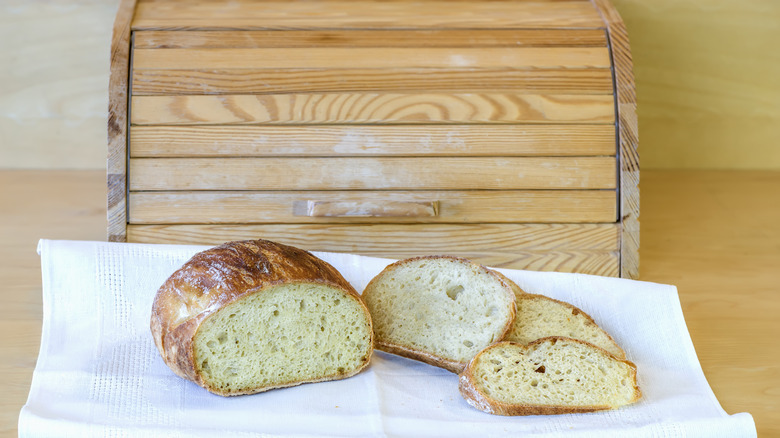 A light wood bread box with a half loaf and slices of bread on a white tea towel on the counter.