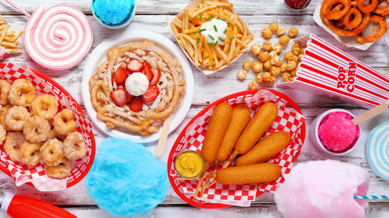 Assorted carnival foods on white wood table including cotton candy, corn dogs, funnel cake, kettle corn, French fries, snow cones, and onion rings
