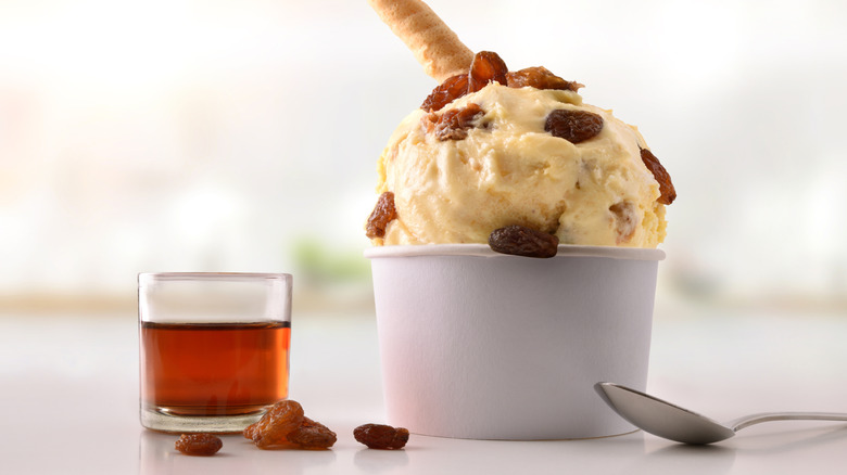 Scoop of rum raisin ice cream in white paper cup next to class of rum, raisins, and silver spoon