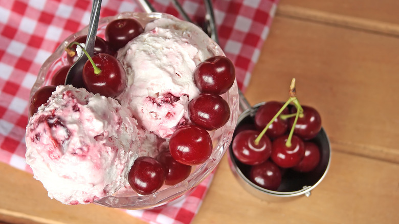 Bowl of two scoops of cherry vanilla ice cream garnished with fresh cherries next to bowl of cherries and red checkered cloth