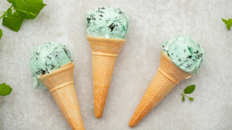 Three mint chocolate chip ice cream cones on gray surface next to mint leaves