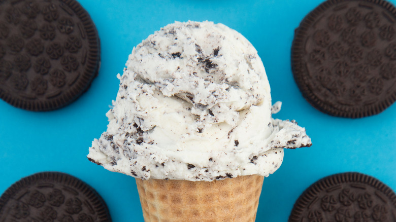 Cookies and cream ice cream in cone against blue background with whole chocolate cookies