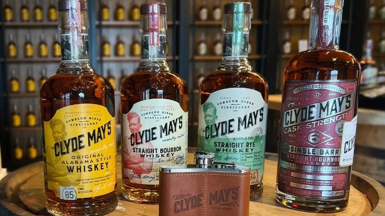 A suite of Clyde May's whiskey bottles on a barrel in a retail shop with a flask in the foreground.