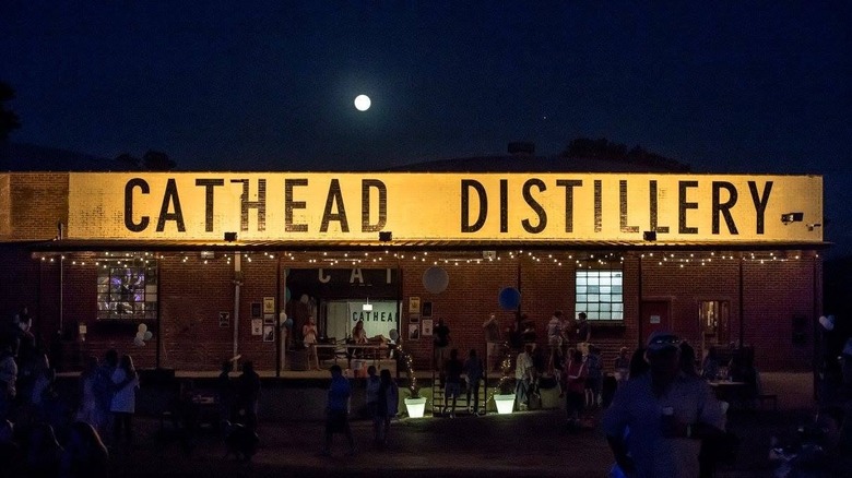 An external shot of Cathead Distillery, a one story brick warehouse building, under full moon with people gathered outside.