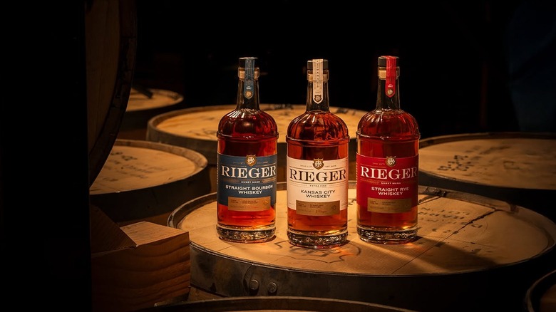 Three different expressions of Rieger whiskeys in glass bottles sitting atop a whiskey barrel in dim light.