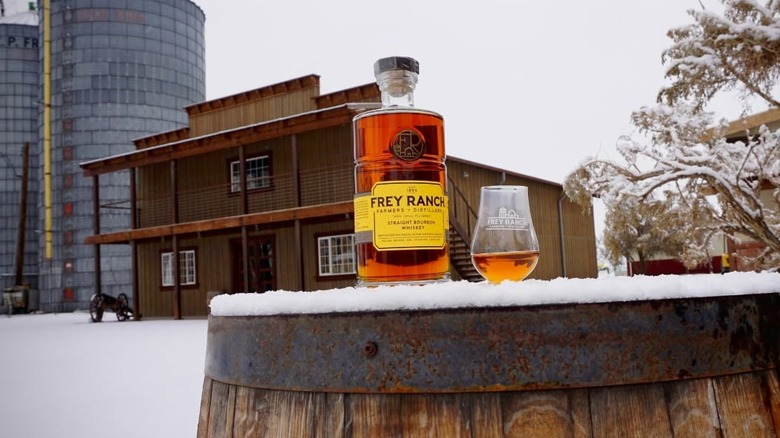 A bottle of Frey Ranch whiskey alongside a snifter glass pour on a snow-covered barrel outside a saloon-style building with silos in the background.