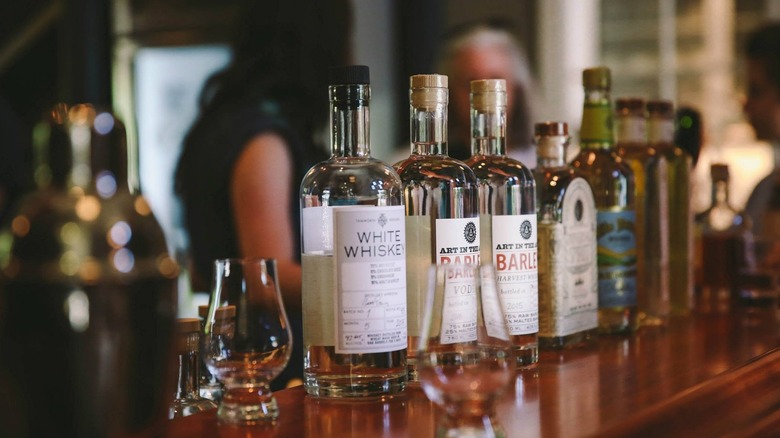 Bottle of Tamworth White Whiskey next to other brands in clear glass bottles and snifters on a wooden bar with people in the background.