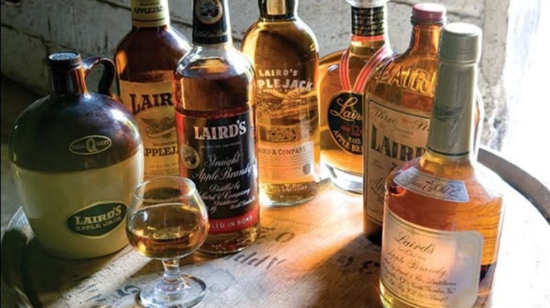 A variety of Laird's Applejack and brandy bottles alongside snifter pour and ceramic jug on a whiskey barrel.