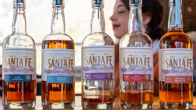 Five expressions of Santa Fe Whiskey in bottles with speed-pour tops with a woman in the background.