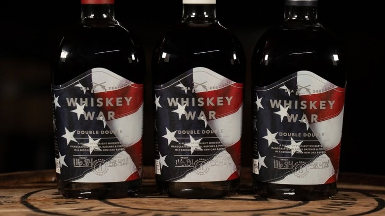 A close-up shot of Whiskey War label. Three bottles white red, white, and blue caps on a barrel against black background.