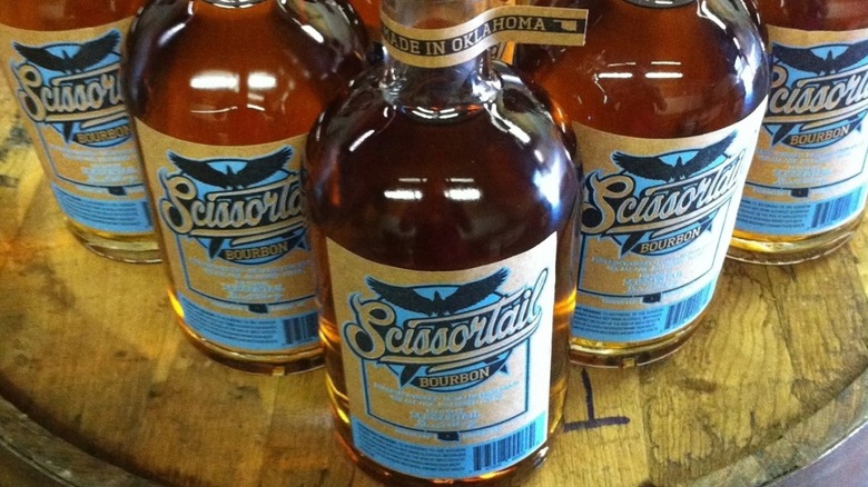A close-up shot of a flying V formation of ScissorTail bourbon bottles with "Made In Oklahoma" tag on neck sitting atop a whiskey barrel with