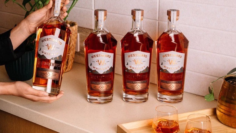 Three bottles of Westward Whiskey in a row with one displayed in a woman's hands. In the foreground sit two pours of whiskey on a cutting board.