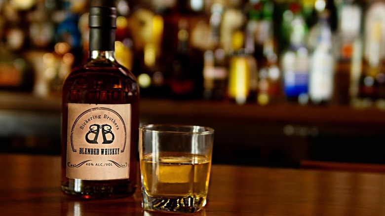 A bottle of Blended Whiskey next to a shot glass pour with a blurry bar in the background.