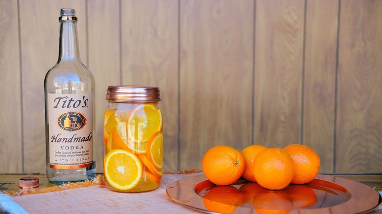 An open bottle of Tit's next to a mason jar of sliced oranges and whole fruit on a copper tray on a table.