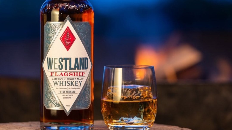 Close-up shot of Westland flagship whiskey bottle next to pour over ice with a campfire in the background.