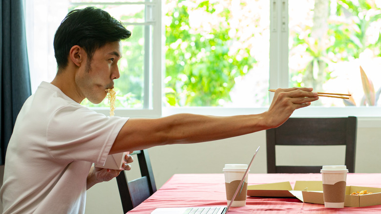 A man with noodles in his mouth pointing across a table with chopsticks