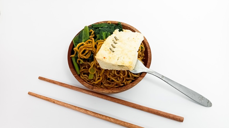 A piece of tofu on a fork resting on a bowl of noodles
