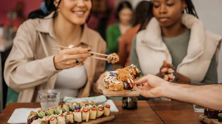 A woman using chopsticks to take sushi off a plate being handed to her