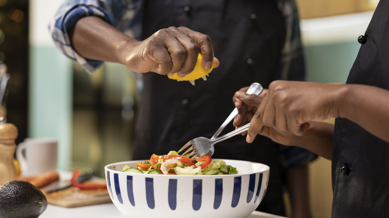 Two people preparing a salad together--one person is tossing the salad while another squeezes a half of a lemon over the bowl