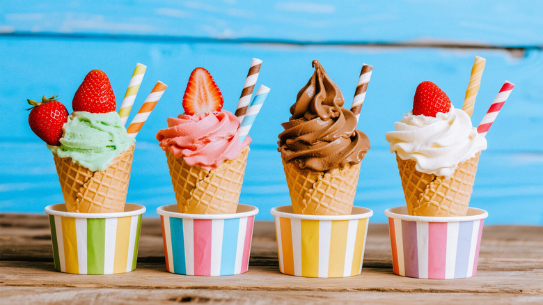 Various flavors of ice cream cones in cups with strawberry and striped straw garnishes against a light blue background.