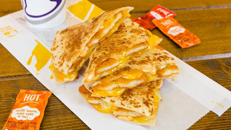 a Taco Bell quesadilla artfully stacked on its paper sleeve on a wooden table with sauce packets and a drink beside it