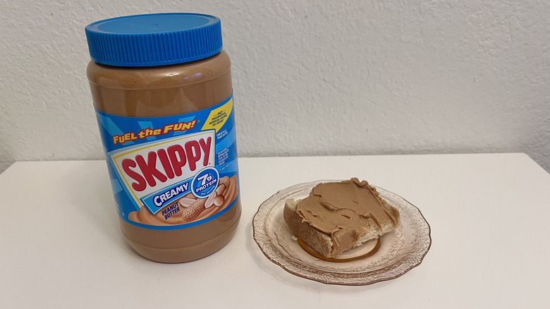 Skippy peanut butter jar beside a piece of toast with the spread on it, on a white table
