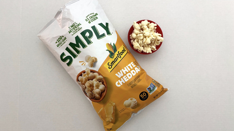 Bag of Smartfood Simply White Cheddar popcorn on white surface next to small bowl of popcorn