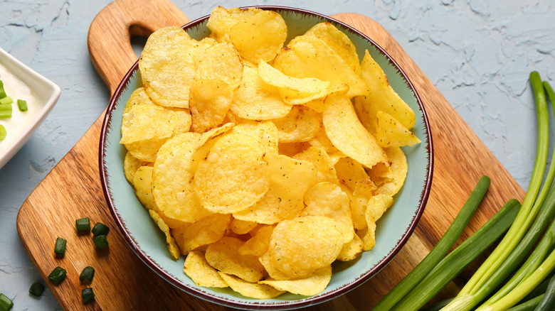 Top view of a bowl full of potato chips, placed on a wooden cutting board alongside spring onions.