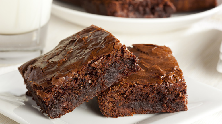 A pair of square cut, dark, chocolatey brownies with a crusty top sitting together on a plate