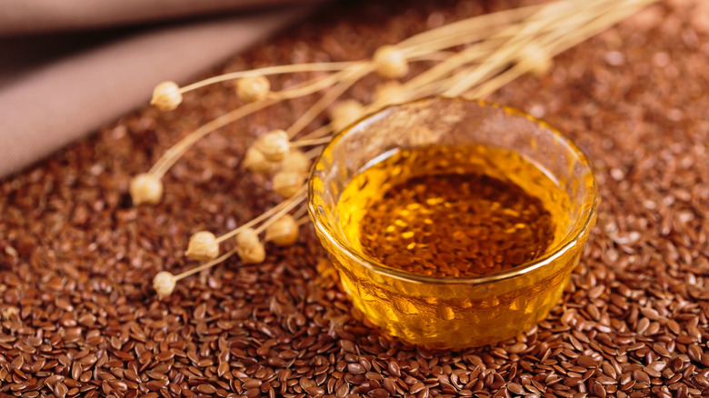 Small bowl of flaxseed oil resting on a surface sprinkled with natural flax seeds, with dried flax seed stalks in the background