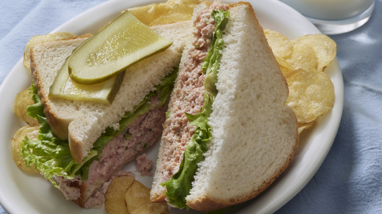 A deviled ham sandwich on a white plate, accompanied by potato chips and two slices of thinly sliced pickle