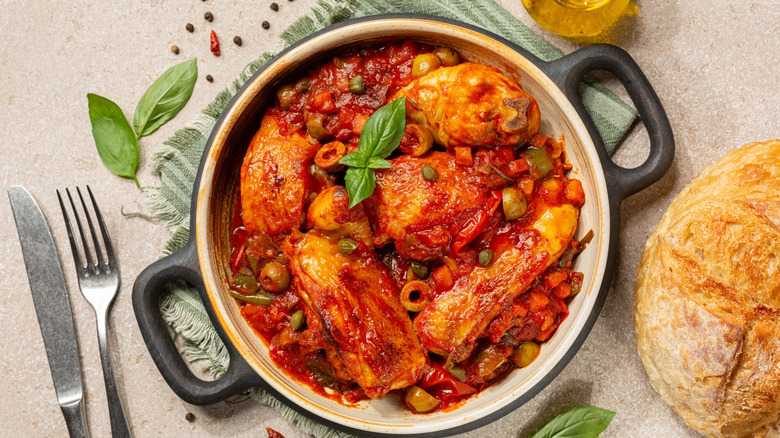 A silver knife and fork are on a beige laminate countertop next to a striped green towel with a pot of chicken cacciatore on top, showing the detailed ingredients like olives, peas, and chicken, served next to a loaf of rustic bread