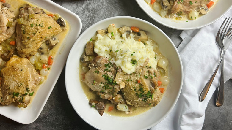 A white plate with a brown-colored rim contains chicken in a creamy sauce with corn, green onions, mushrooms, and tomatoes and a side of rice.