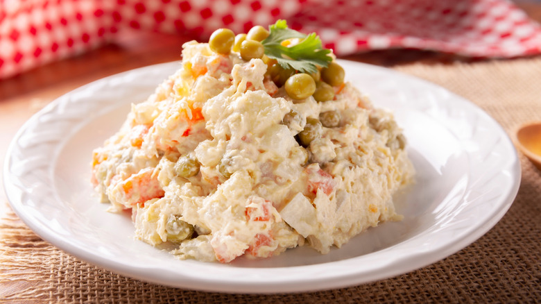 A white plate on a natural fiber placemat in front of a red and white checkered tablecloth is a white bowl plate served with a heaping pile of Russian or Olivier salad shows the detailed chicken, potatoes, and vegetable salad, topped with green peas