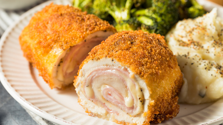 A closeup of breaded chicken sliced in half to show it's stuffed with swirls of ham and cheese on a white plate, served with broccoli and mashed potatoes topped with a heavy seasoning