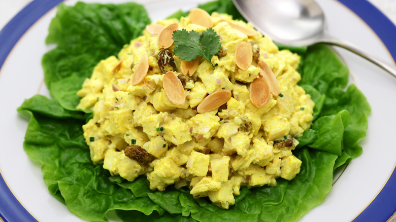 A bed of lettuce on a white plate with blue trim is topped with an organized pile of cubed yellow curry chicken salad with almonds sprinkled on top