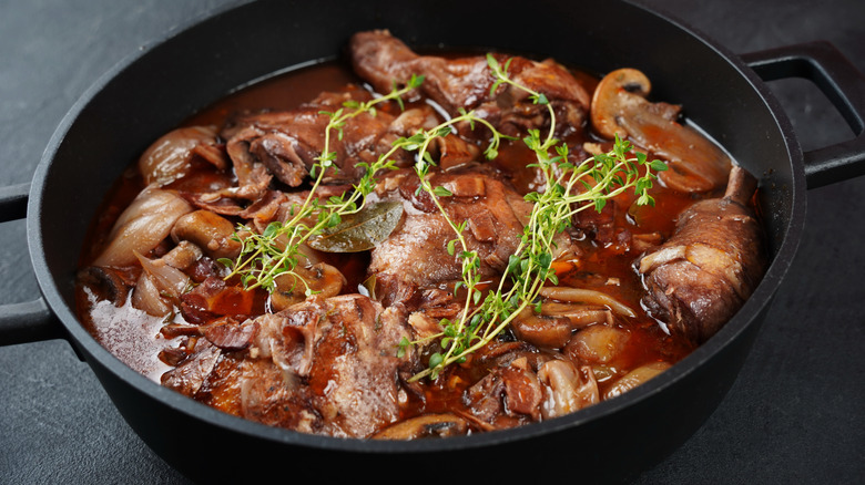 A black cast iron skillet is filled with a dark red wine broth and rooster meat, topped with microgreens