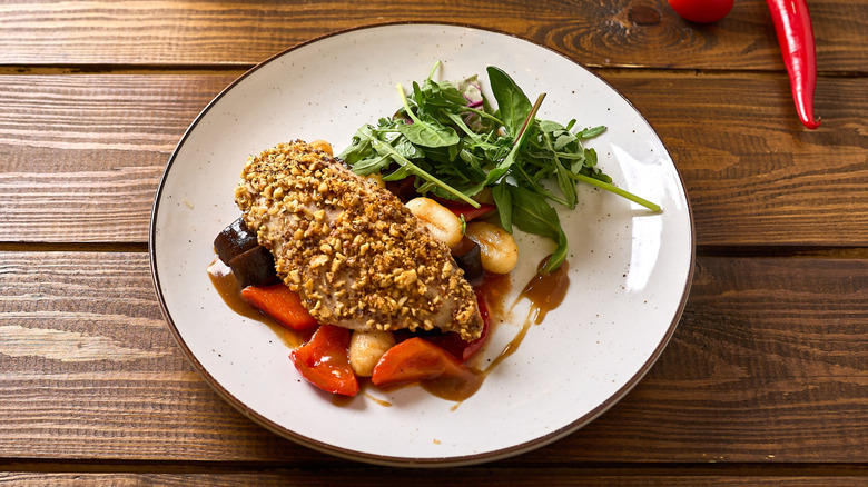 A wooden table is topped with a white plate that is filled with almond-crusted chicken, roasted vegetables, fresh arugula, and a red-wine glaze drizzled on top. A red chili pepper is visible in the top right corner of the photo.