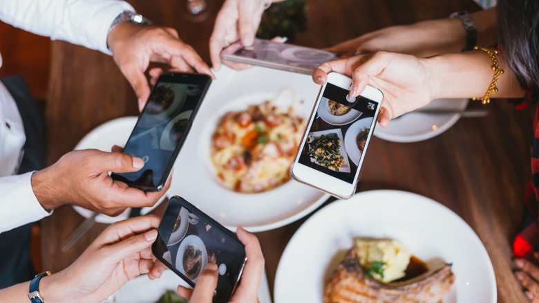 Four people holding their phones up at a table to take pictures of food at a restaurant.