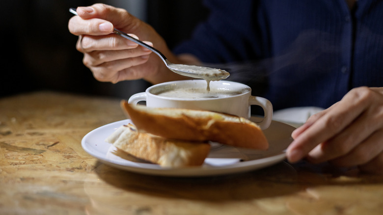 A person eats soup from a white bowl with bread on the plate