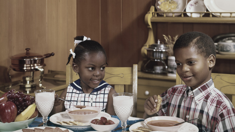 A boy and a girl eat a soup at a full table, circa 1960