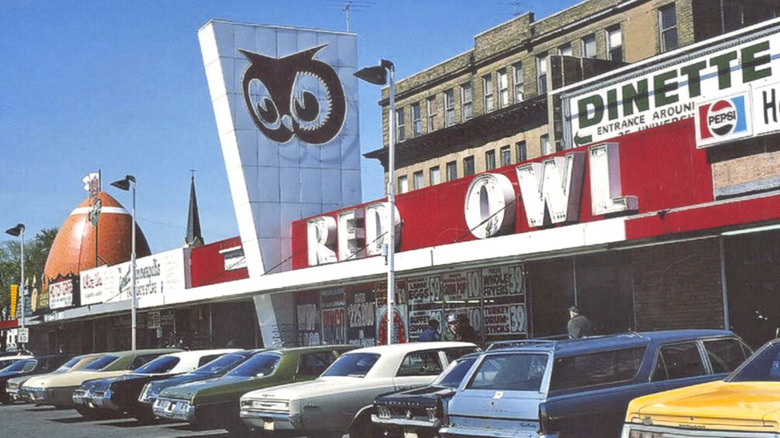 Vintage photograph showing exterior of Red Owl grocery store with red banner, large logo sign, and cars parked outside