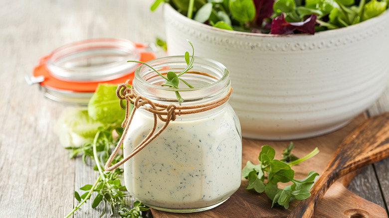 A phyto jar of creamy, herb flecked dressing sits on a cutting board, with a bowl of salad in the background