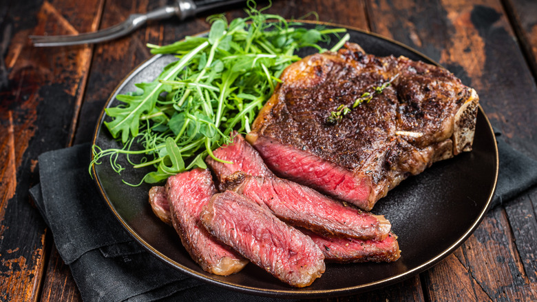 A partially sliced steak on a black plate accompanied by an arugula salad
