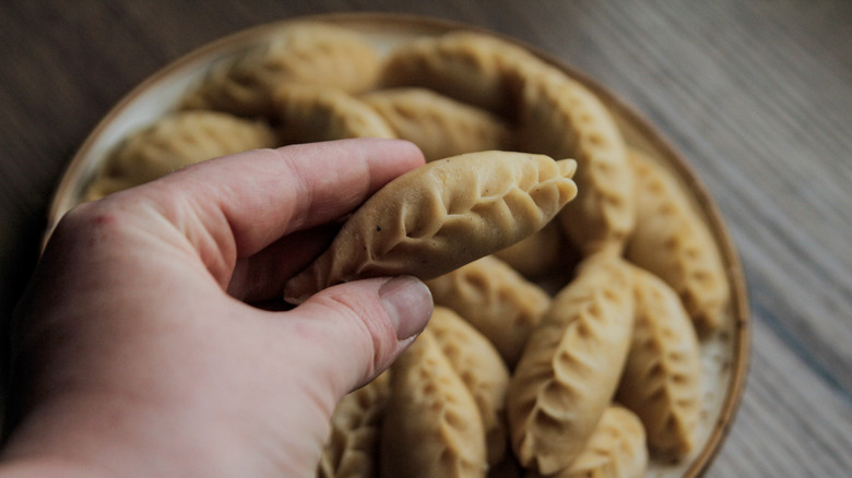Someone's hand holding a freshly made culurgione stuffed pasta in a wheat spike shape above a bowl of culurgioni