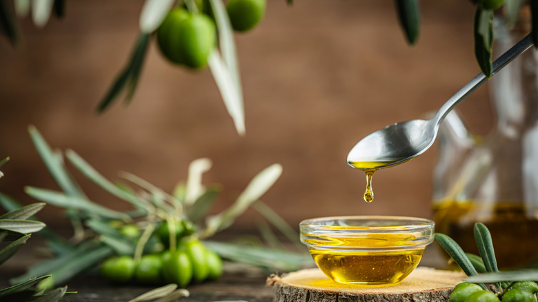 A spoon drips olive oil into a small glass bowl wth olive branches in the background.