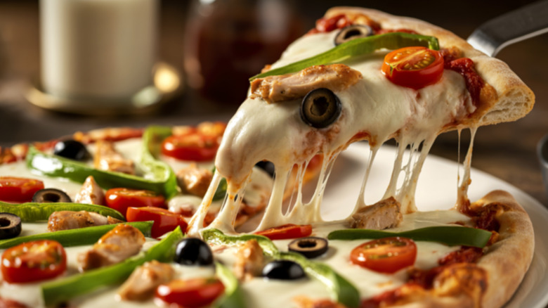 Someone lifting a slice of cheesy pizza covered in olives, peppers, tomatoes, and chicken