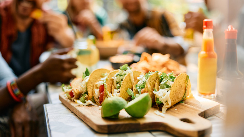 A board of tacos and sliced limes with customers laughing in the background